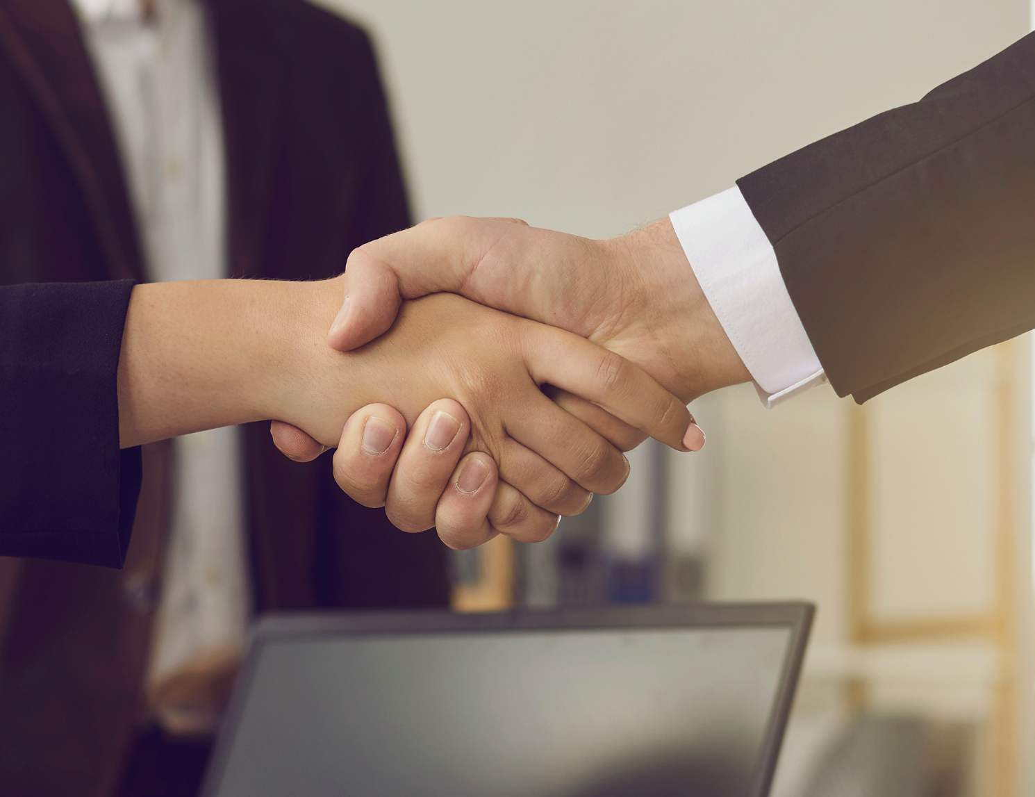Two business professionals shake hands across a desk, symbolising a successful partnership or agreement. A laptop, notebook and office workspace are visible in the foreground, while another person stands blurred in the background.